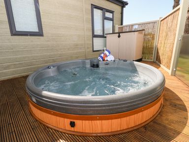 An outdoor wooden deck with a round hot tub next to a building with windows and a storage box with towels and a bottle at Beechwood Lodge in Hasguard Cross near Broad Haven