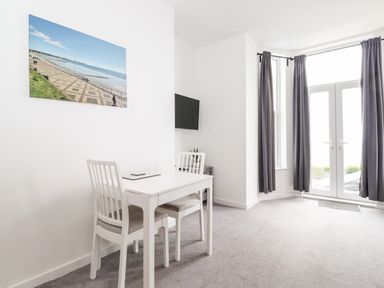 A dining area with a white table and two chairs next to a wall-mounted TV and large windows with curtains at 3 Seaview @ Bridlington Bay in Bridlington