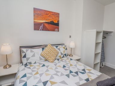 A bedroom with a bed with geometric patterned bedding two bedside lamps a white open wardrobe with hanging space and shelves and a sunset photograph on the wall at 4 Seaview Bridlington Bay in Bridlington