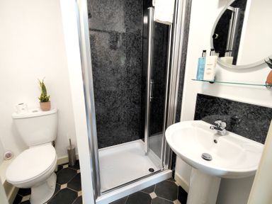 A bathroom with a toilet, a shower with black walls, a sink, and a round mirror at The Paddock in Keswick