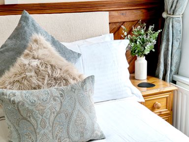 A bed with multiple decorative pillows next to a wooden nightstand with a vase of flowers and curtains at The Paddock in Keswick