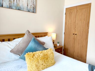 A bedroom with a wooden bed frame and cushions beside a wooden closet and a bedside table with a lamp and plant at The Paddock in Keswick