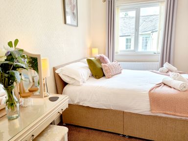 A bedroom with a bed with pillows and rolled towels next to a window and a vanity with flowers and a lamp at The Paddock in Keswick