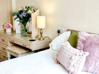 A bedroom with a bed with decorative pillows next to a dressing table with a mirror lamp flowers and tissues at The Paddock in Keswick