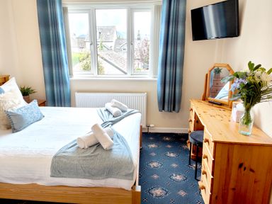 A bedroom with a bed, blue curtains, a wooden dresser with a mirror, and a wall-mounted TV at The Paddock in Keswick