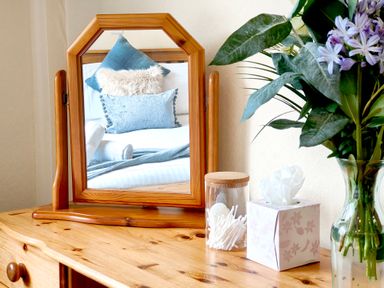 A wooden dressing table with a mirror reflecting a bed with pillows next to a jar of cotton swabs a tissue box and a vase with flowers at The Paddock in Keswick