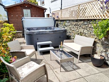 An outdoor patio with wicker chairs a loveseat a glass top table a hot tub a wooden shed and stone wall at The Paddock in Keswick