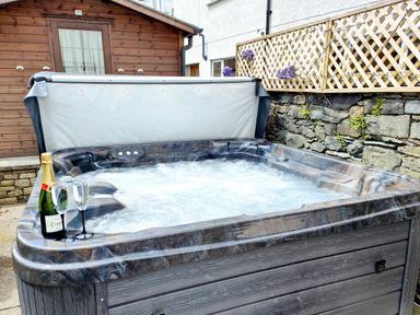 An outdoor hot tub with bubbling water a bottle of champagne and two glasses next to a wooden shed and a stone wall with a lattice fence at The Paddock in Keswick