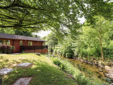 A wooden lodge next to a stream surrounded by grass and trees at Nutkins Lodge Beckside 5 Limefitt Park near Windermere
