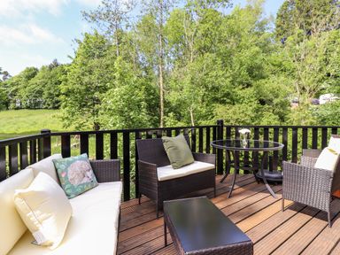 A wooden deck with wicker furniture and cushions overlooking trees and greenery at Nutkins Lodge Beckside 5 Limefitt Park near Windermere