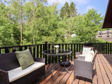 A wooden deck with wicker chairs and a glass table overlooking trees at Nutkins Lodge in Beckside 5 Limefitt Park near Windermere