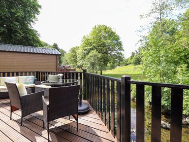 An outdoor deck with wicker chairs a glass table and a sofa overlooking a grassy field and trees at Nutkins Lodge in Beckside 5 Limefitt Park near Windermere