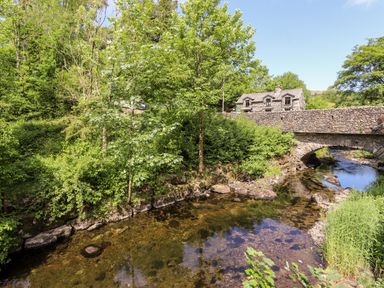 A stone bridge over a river with trees and a stone house in the background at Nutkins Lodge Beckside 5 Limefitt Park near Windermere