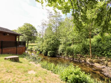 A wooden cabin next to a small stream surrounded by trees and grass at Nutkins Lodge Beckside 5 Limefitt Park near Windermere