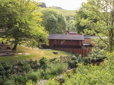 A wooden cabin with a fenced porch surrounded by trees and greenery near a small stream at Nutkins Lodge in Beckside 5 Limefitt Park near Windermere