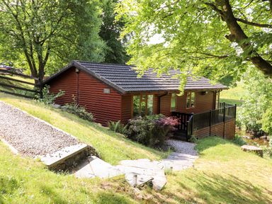 A wooden lodge with a black tiled roof surrounded by trees and greenery at Nutkins Lodge Beckside 5 Limefitt Park near Windermere