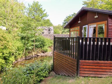 A wooden cabin with a black railing next to a clear stream surrounded by trees at Nutkins Lodge Beckside 5 Limefitt Park near Windermere