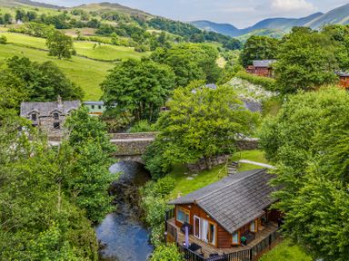 A wooden cabin near a river with a stone bridge and green hills in the background at Nutkins Lodge Beckside 5 Limefitt Park near Windermere