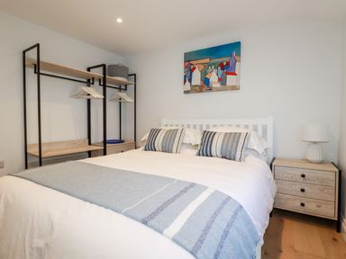 A bedroom with a double bed pillows a nightstand with a lamp an open wardrobe with hangers and a painting on the wall at Beach Cottage in Cadgwith