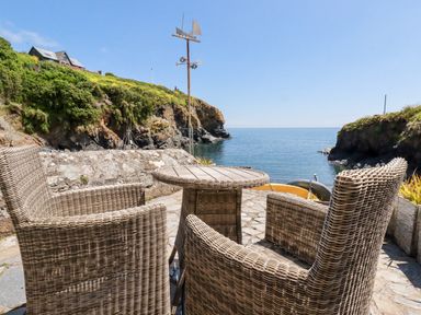 A patio with wicker chairs and a table overlooking a rocky coastline and the sea at Beach Cottage in Cadgwith