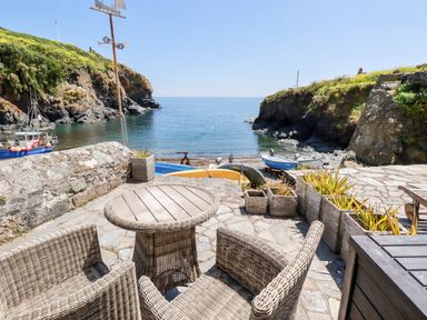 An outdoor stone patio with a round table and wicker chairs overlooking boats and the sea at Beach Cottage in Cadgwith