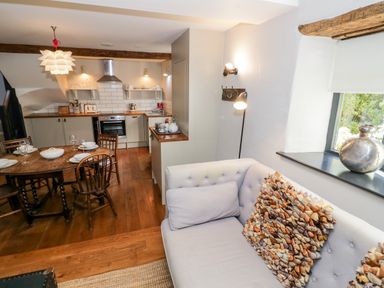 A kitchen and dining area with a round wooden table and chairs and a sofa with cushions near a window at The Old Stables in Chipping Campden