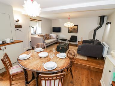 A dining area with a round wooden table and four chairs set with dishes adjacent to a living room with two sofas a black chair a black stove and a TV at The Old Stables in Chipping Campden