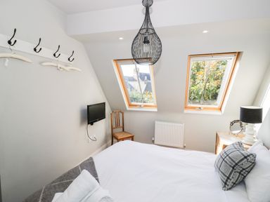 A bedroom with a bed pillow and blanket a chair a wall-mounted TV two windows and a side table with a lamp at The Old Stables in Chipping Campden