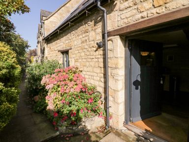 A stone exterior wall with pink flowers and a partially open dark door at The Old Stables in Chipping Campden