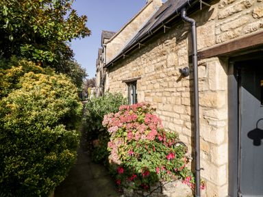 A stone exterior wall with a black door and window next to a narrow garden path with bushes and flowering plants at The Old Stables in Chipping Campden