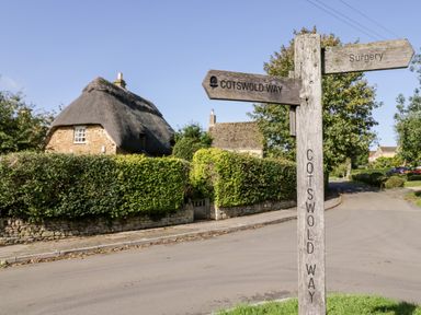 A street intersection with a wooden signpost indicating Cotswold Way and Surgery near a thatched roof cottage at The Old Stables in Chipping Campden