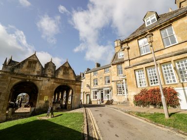 A street with historic stone buildings and an archway structure under a partly cloudy sky at The Old Stables in Chipping Campden
