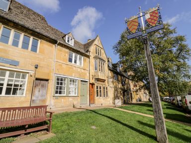 A row of stone buildings with windows and doors and a wooden bench on grass near a signpost in Chipping Campden