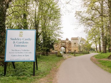 A road leading to an entrance gate with a stone building behind and a large sign for Sudeley Castle and Gardens Entrance