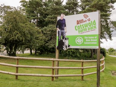 An outdoor sign for Adam Henson's Cotswold Farm Park beside a wooden fence and trees