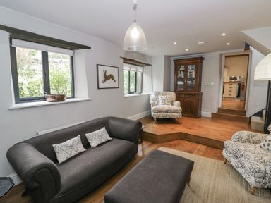 A living room with a dark grey sofa, two patterned armchairs, wooden cabinet, and windows at The Old Brewhouse in Chipping Campden