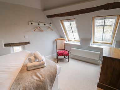 A bedroom with a bed, two windows, a wooden chair with a red cushion, a chest of drawers and coat hooks on the wall at The Old Brewhouse in Chipping Campden