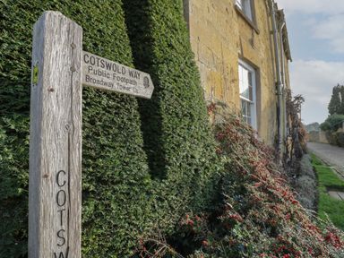 A wooden signpost with Cotswold Way public footpath directions near a stone building and hedges at The Old Brewhouse in Chipping Campden