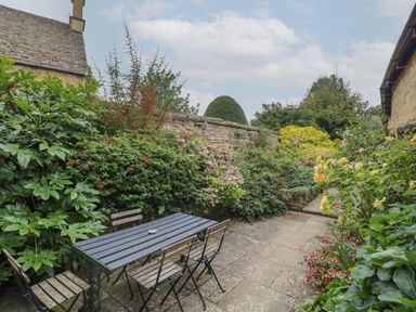 An outdoor patio with a metal table and wooden chairs surrounded by various green plants and flowers at The Old Brewhouse in Chipping Campden