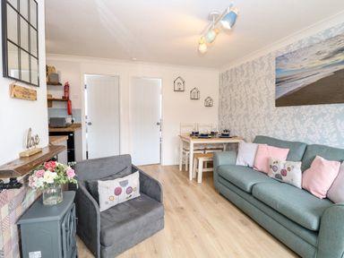 A living area with a grey armchair and a teal sofa with cushions a wooden dining table set against a wall with leaf pattern wallpaper at 72 in Hemsby