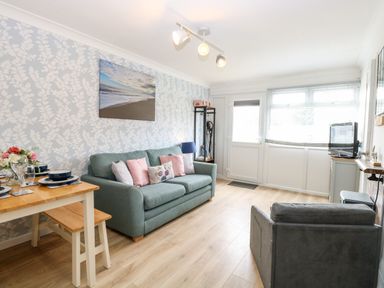 A living room with a green sofa, a grey armchair, dining table with bench and plates, and a wall with leaf patterned wallpaper at 72 in Hemsby