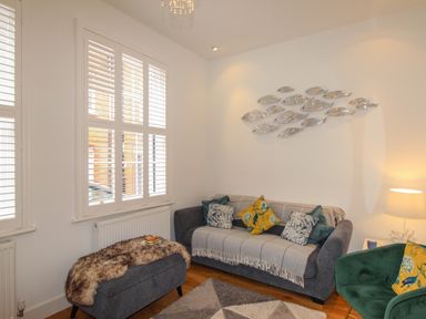 A living room with a grey sofa covered by a throw with cushions a green armchair a grey padded bench with a fur throw and metal fish wall decor at The Cottage in Weymouth
