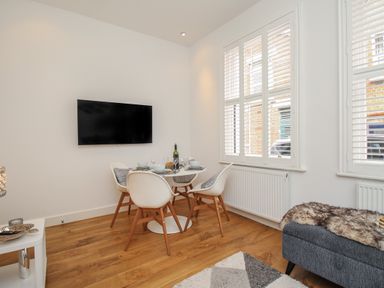 A dining area with a round table set for four and a wall-mounted television in a room with large windows at The Cottage in Weymouth