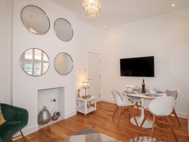 A dining area with a round table set for four and a wall-mounted TV with mirrors on the adjacent wall at The Cottage in Weymouth