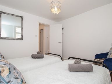 A bedroom with two beds each having a rolled towel on top a blue armchair a large mirror on the wall and an open door at The Cottage in Weymouth