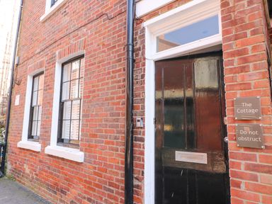 A black front door with two signs next to it on a brick building with two windows at The Cottage in Weymouth
