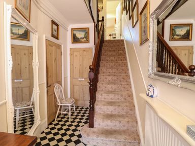 A hallway with a carpeted staircase a white chair checkered floor wooden doors and framed pictures at The Studio in Sheringham