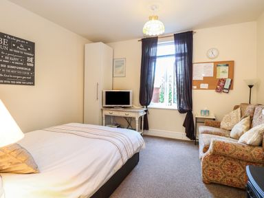 A bedroom with a bed sofa television on a white table and a window with dark curtains at The Studio in Sheringham