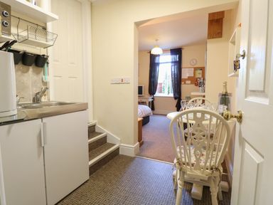 A kitchenette with a sink and cabinets next to a small dining table and chairs leading into a bedroom with a window and a bed at The Studio in Sheringham