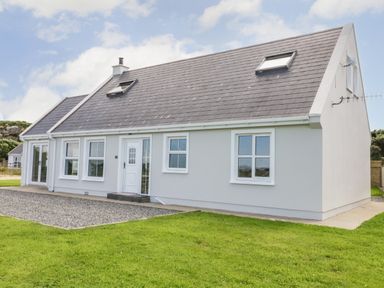 A single story house with grey walls, a dark grey roof, white framed windows and a white door surrounded by grass at The Haven in Falcarragh County Donegal
